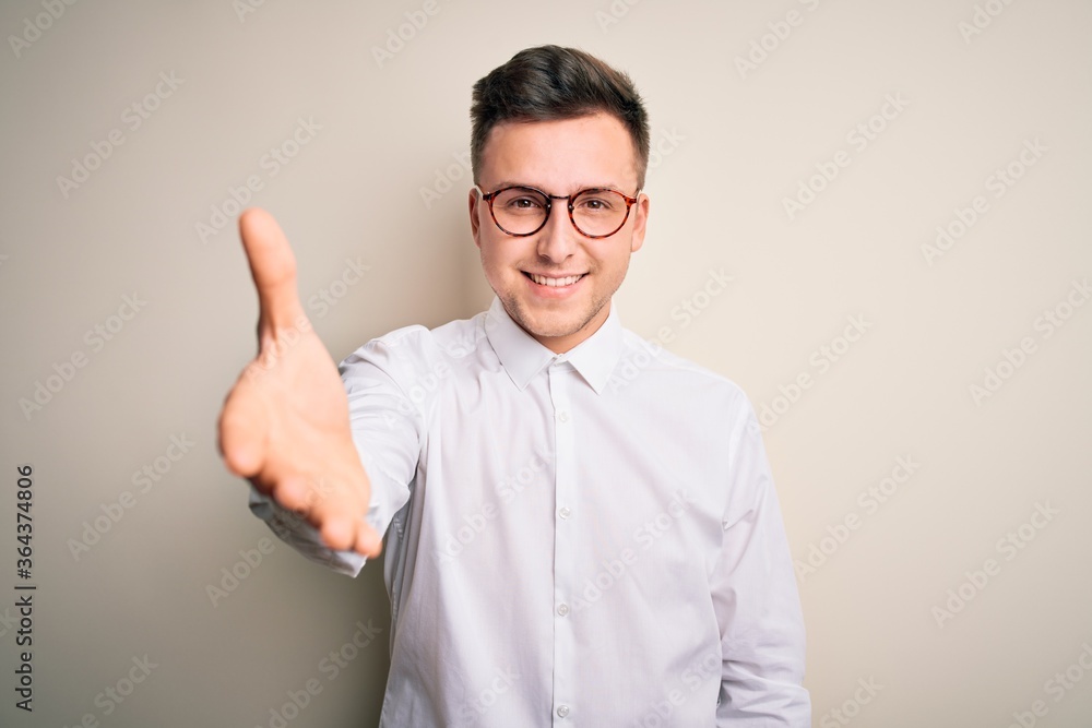 Young handsome business mas wearing glasses and elegant shirt over isolated background smiling friendly offering handshake as greeting and welcoming. Successful business.