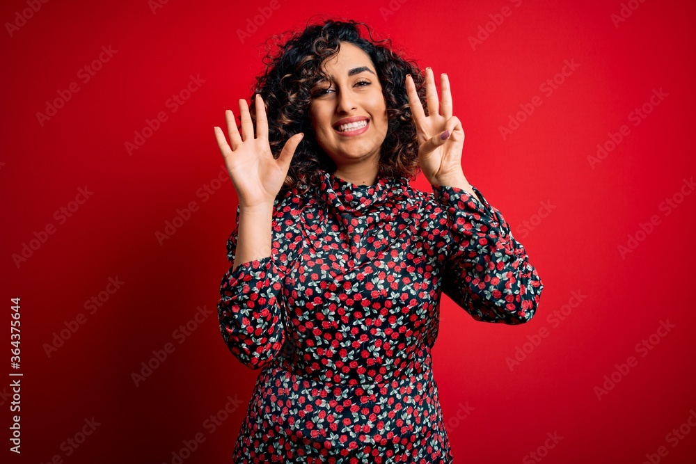 Young beautiful curly arab woman wearing casual floral dress standing over red background showing and pointing up with fingers number eight while smiling confident and happy. Young beautiful curly arab woman wearing casual floral dress standing over red background showing and pointing up with fingers number eight while smiling confident and happy.