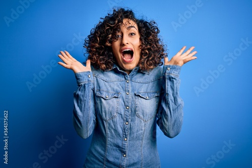 Young beautiful curly arab woman wearing casual denim shirt standing over blue background celebrating crazy and amazed for success with arms raised and open eyes screaming excited. Winner concept