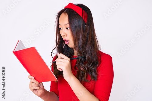 Young beautiful chinese girl holding book scared and amazed with open mouth for surprise, disbelief face