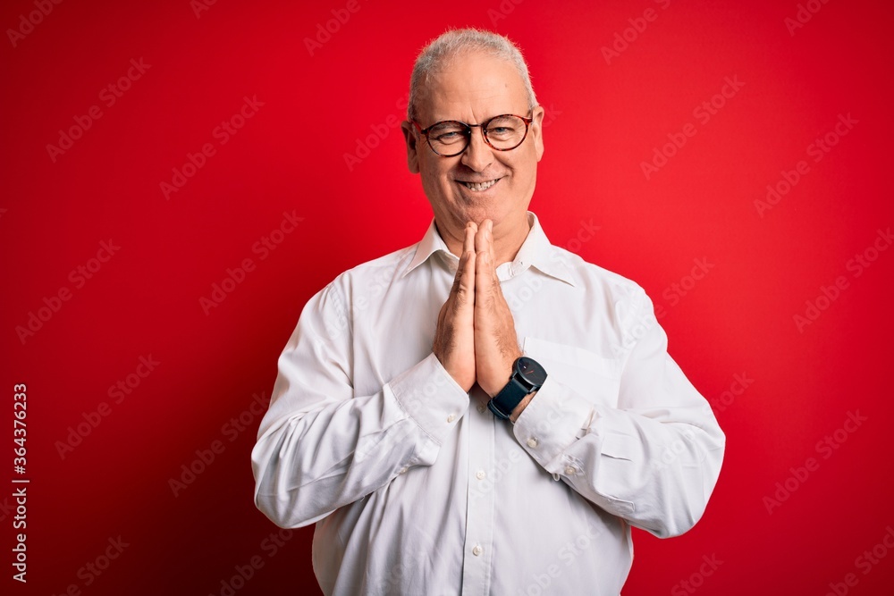 Middle age handsome hoary man wearing casual shirt and glasses over red background praying with hands together asking for forgiveness smiling confident. Middle age handsome hoary man wearing casual shirt and glasses over red background praying with hands together asking for forgiveness smiling confident.