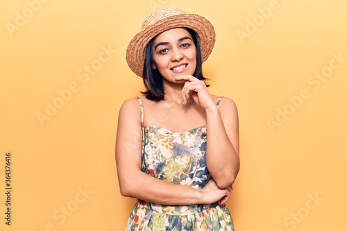 Young beautiful latin woman wearing summer hat looking confident at the camera with smile with crossed arms and hand raised on chin. thinking positive.