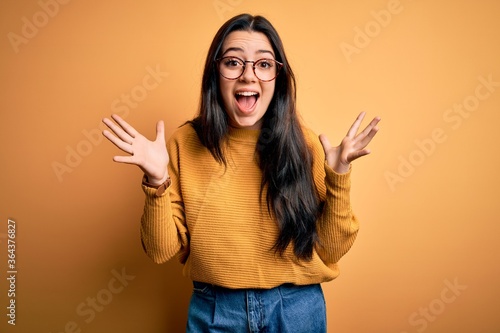 Tableau sur toile Young brunette woman wearing glasses and casual sweater over yellow isolated background celebrating crazy and amazed for success with arms raised and open eyes screaming excited