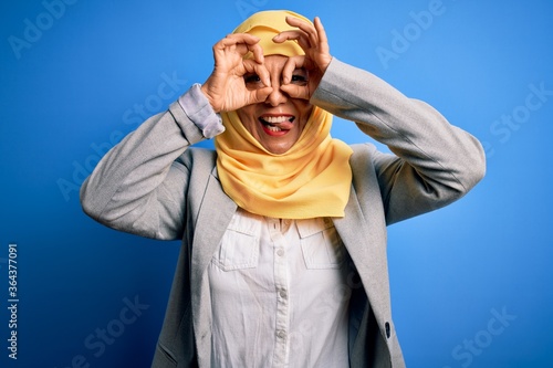 Middle age brunette business woman wearing muslim traditional hijab over blue background doing ok gesture like binoculars sticking tongue out, eyes looking through fingers. Crazy expression.