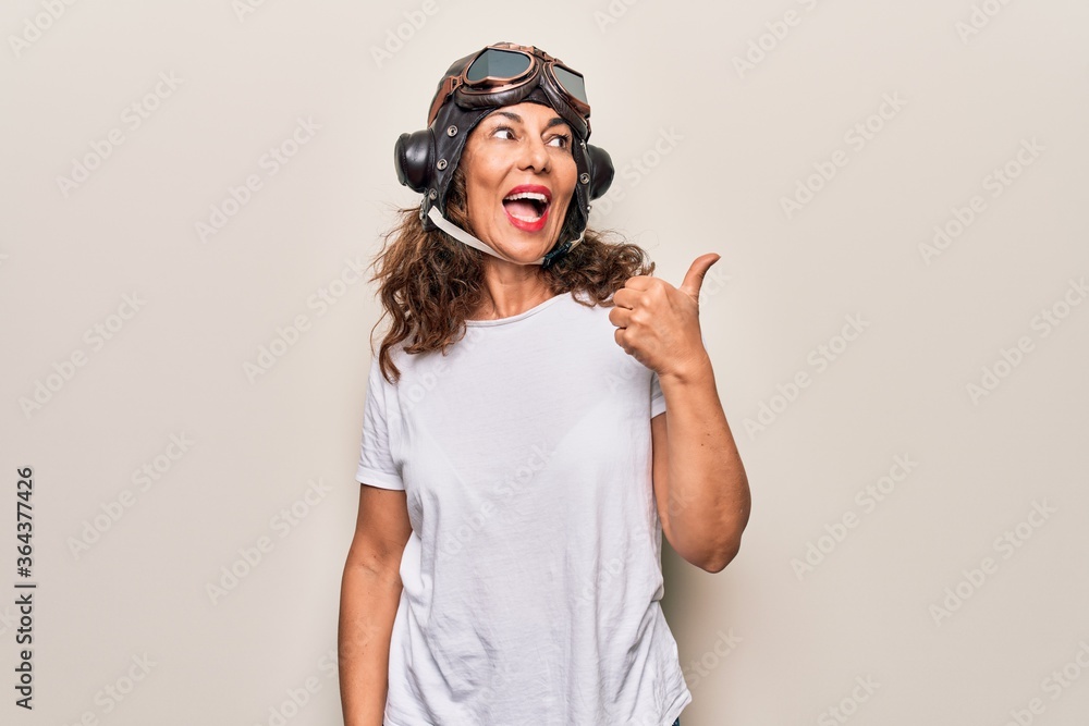 Middle age brunette woman wearing goggles and retro aviator leather hat over white background pointing thumb up to the side smiling happy with open mouth Middle age brunette woman wearing goggles and retro aviator leather hat over white background pointing thumb up to the side smiling happy with open mouth