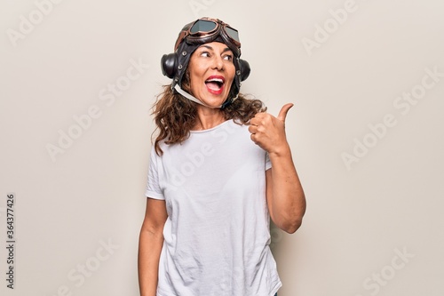 Middle age brunette woman wearing goggles and retro aviator leather hat over white background pointing thumb up to the side smiling happy with open mouth