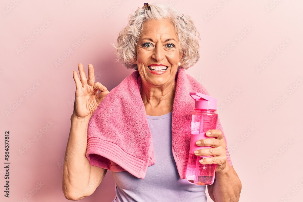 Senior grey-haired woman wearing sportswear and towel drinking bottle of water doing ok sign with fingers, smiling friendly gesturing excellent symbol Senior grey-haired woman wearing sportswear and towel drinking bottle of water doing ok sign with fingers, smiling friendly gesturing excellent symbol