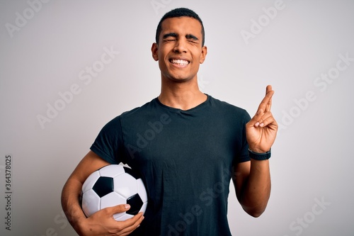 Handsome african american man playing footbal holding soccer ball over white background gesturing finger crossed smiling with hope and eyes closed. Luck and superstitious concept. © Krakenimages.com