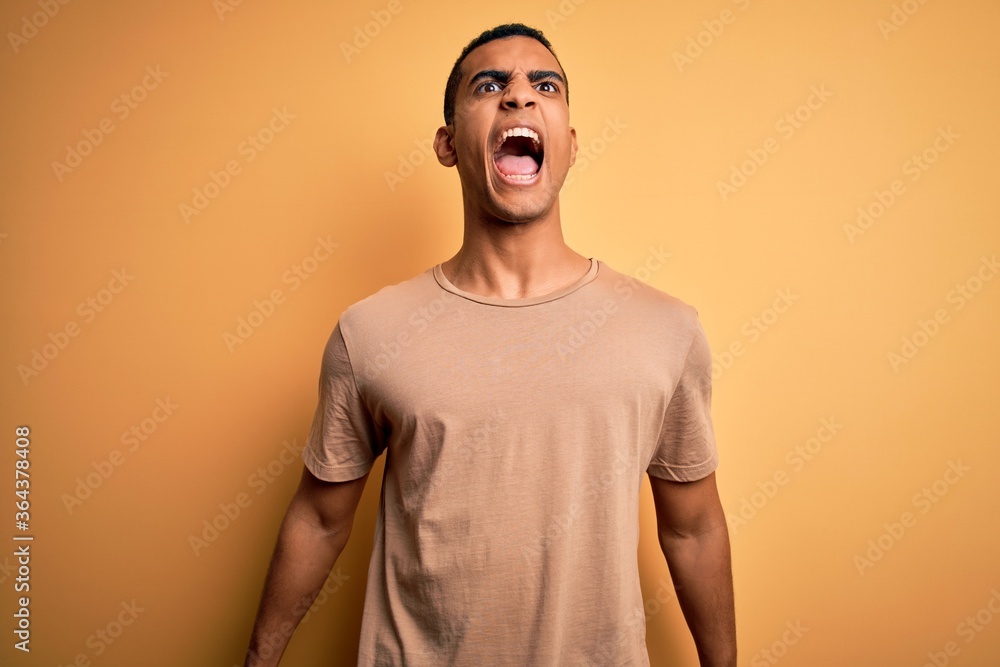 Fototapeta premium Young handsome african american man wearing casual t-shirt standing over yellow background angry and mad screaming frustrated and furious, shouting with anger. Rage and aggressive concept.