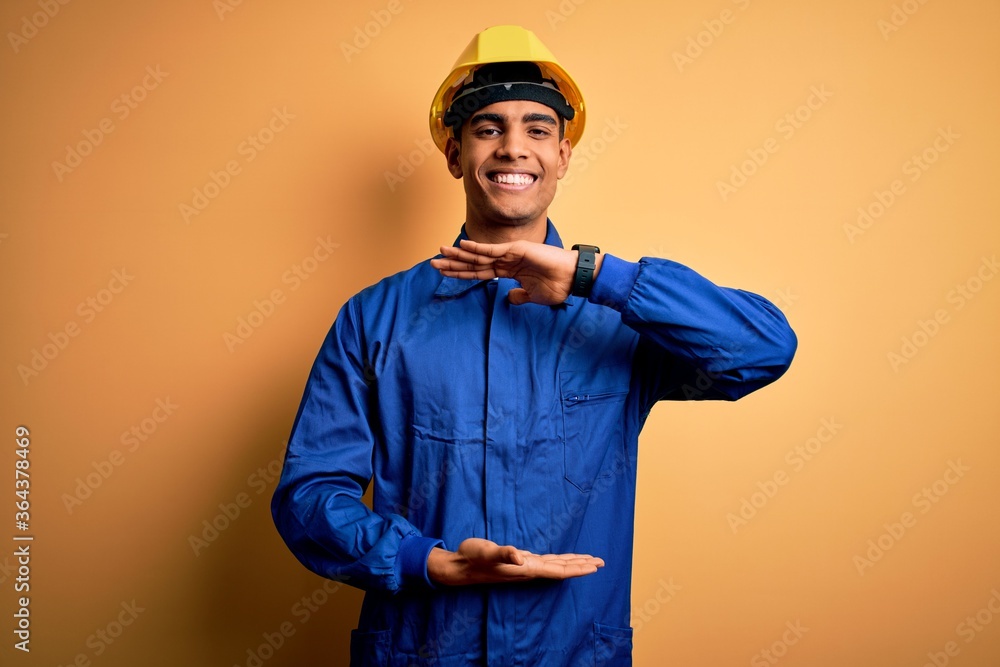 Young handsome african american worker man wearing blue uniform and security helmet gesturing with hands showing big and large size sign, measure symbol. Smiling looking at the camera. Measuring