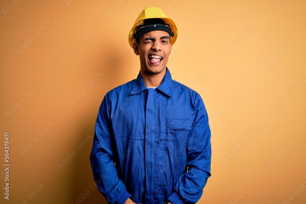 Young handsome african american worker man wearing blue uniform and security helmet winking looking at the camera with sexy expression, cheerful and happy face. Young handsome african american worker man wearing blue uniform and security helmet winking looking at the camera with sexy expression, cheerful and happy face.