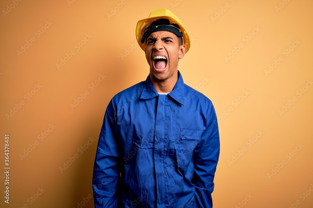 Young handsome african american worker man wearing blue uniform and security helmet angry and mad screaming frustrated and furious, shouting with anger. Rage and aggressive concept.