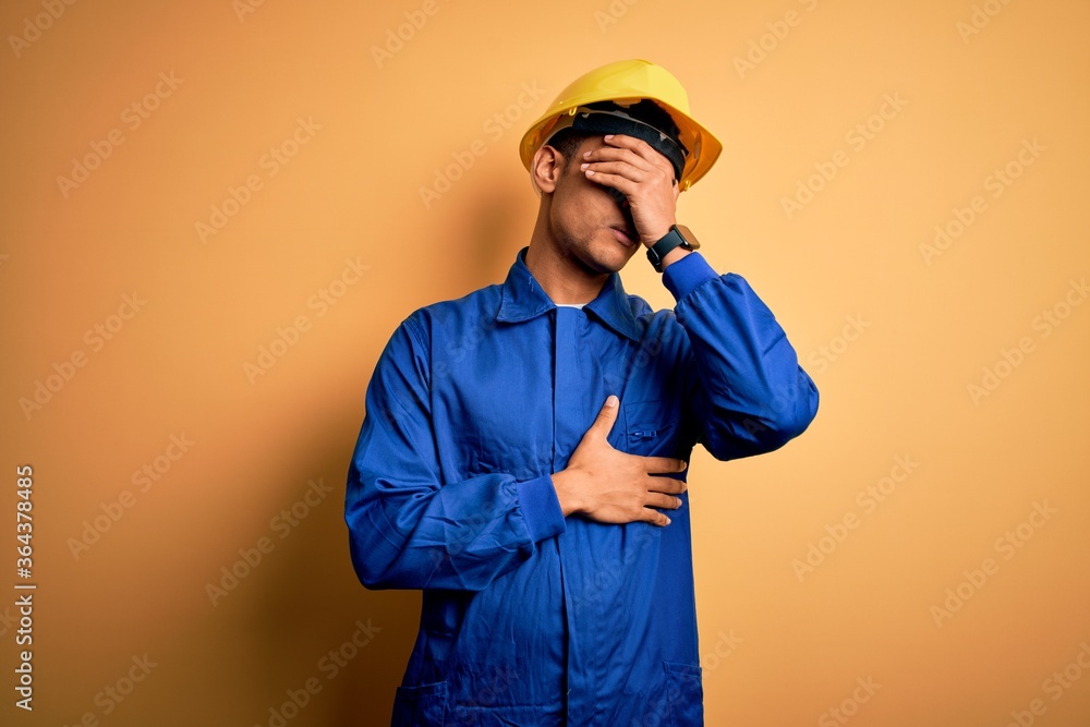 Young handsome african american worker man wearing blue uniform and security helmet Touching forehead for illness and fever, flu and cold, virus sick