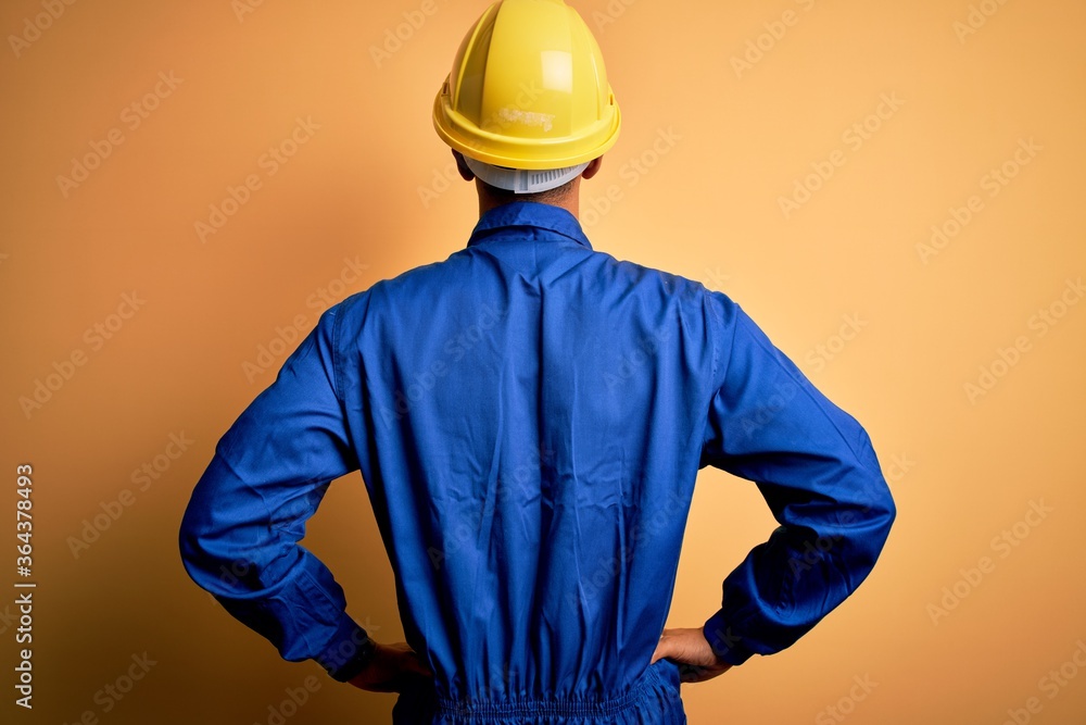 Young handsome african american worker man wearing blue uniform and security helmet standing backwards looking away with arms on body