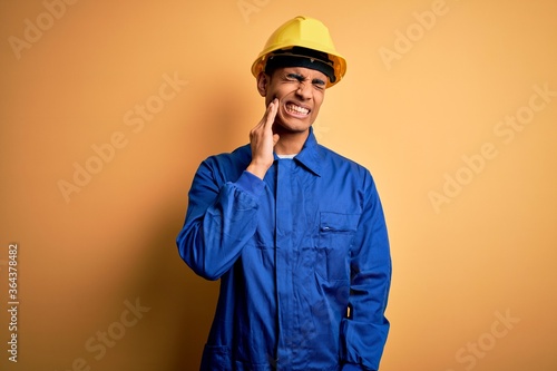 Young handsome african american worker man wearing blue uniform and security helmet touching mouth with hand with painful expression because of toothache or dental illness on teeth. Dentist