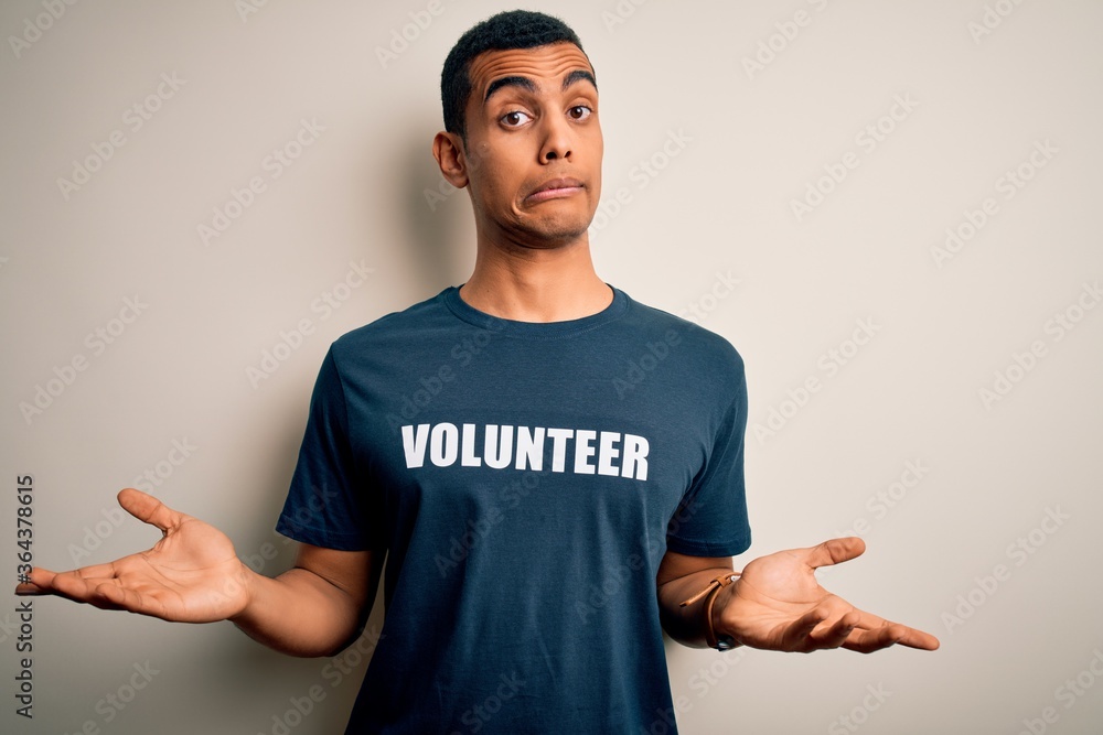 Young handsome african american man volunteering wearing t-shirt with volunteer message clueless and confused expression with arms and hands raised. Doubt concept. Young handsome african american man volunteering wearing t-shirt with volunteer message clueless and confused expression with arms and hands raised. Doubt concept.