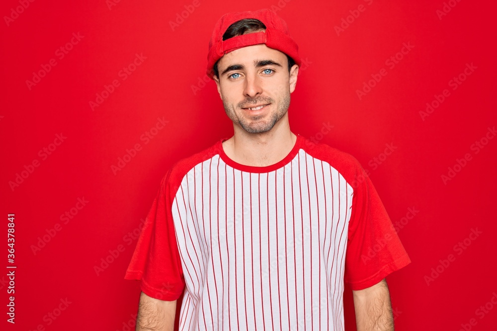 Young handsome sporty man with blue eyes wearing striped baseball t-shirt and cap with a happy and cool smile on face. Lucky person.