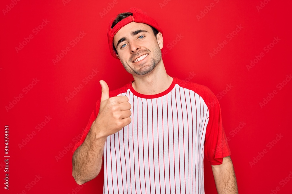 Young handsome sporty man with blue eyes wearing striped baseball t-shirt and cap doing happy thumbs up gesture with hand. Approving expression looking at the camera showing success. Young handsome sporty man with blue eyes wearing striped baseball t-shirt and cap doing happy thumbs up gesture with hand. Approving expression looking at the camera showing success.