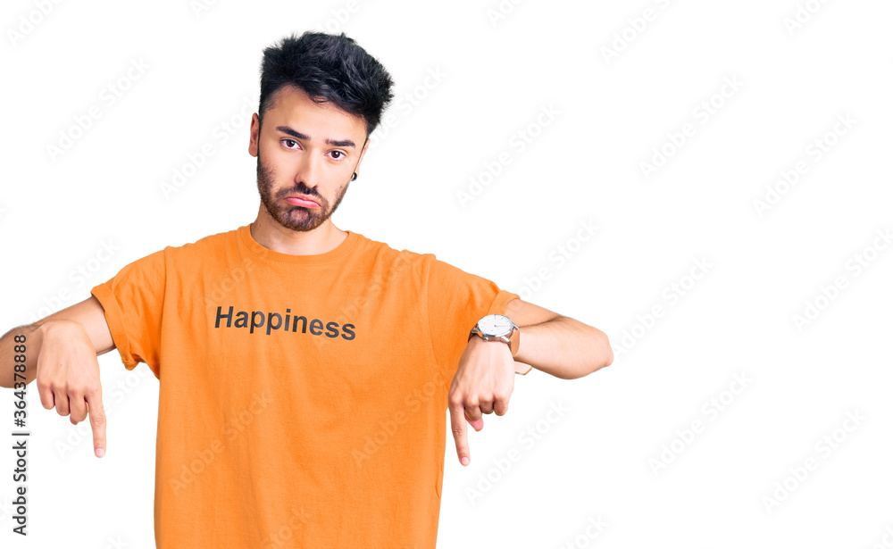 Young hispanic man wearing t shirt with happiness word message pointing down looking sad and upset, indicating direction with fingers, unhappy and depressed. Young hispanic man wearing t shirt with happiness word message pointing down looking sad and upset, indicating direction with fingers, unhappy and depressed.