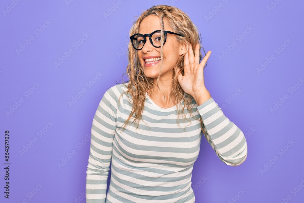 Beautiful blonde woman wearing casual striped t-shirt and glasses over purple background smiling with hand over ear listening an hearing to rumor or gossip. Deafness concept. Beautiful blonde woman wearing casual striped t-shirt and glasses over purple background smiling with hand over ear listening an hearing to rumor or gossip. Deafness concept.