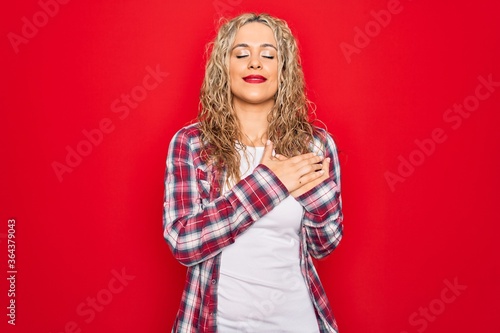 Young beautiful blonde woman wearing casual shirt standing over isolated red background smiling with hands on chest with closed eyes and grateful gesture on face. Health concept.