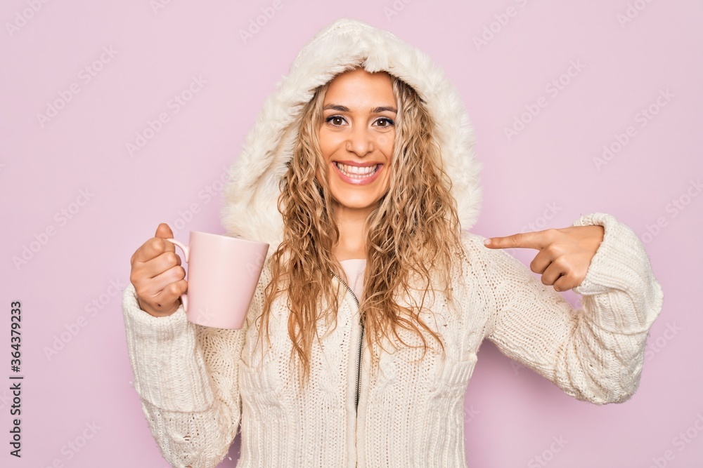 Young beautiful blonde woman wearing casual sweater with hood drinking cup of coffee pointing finger to one self smiling happy and proud Young beautiful blonde woman wearing casual sweater with hood drinking cup of coffee pointing finger to one self smiling happy and proud