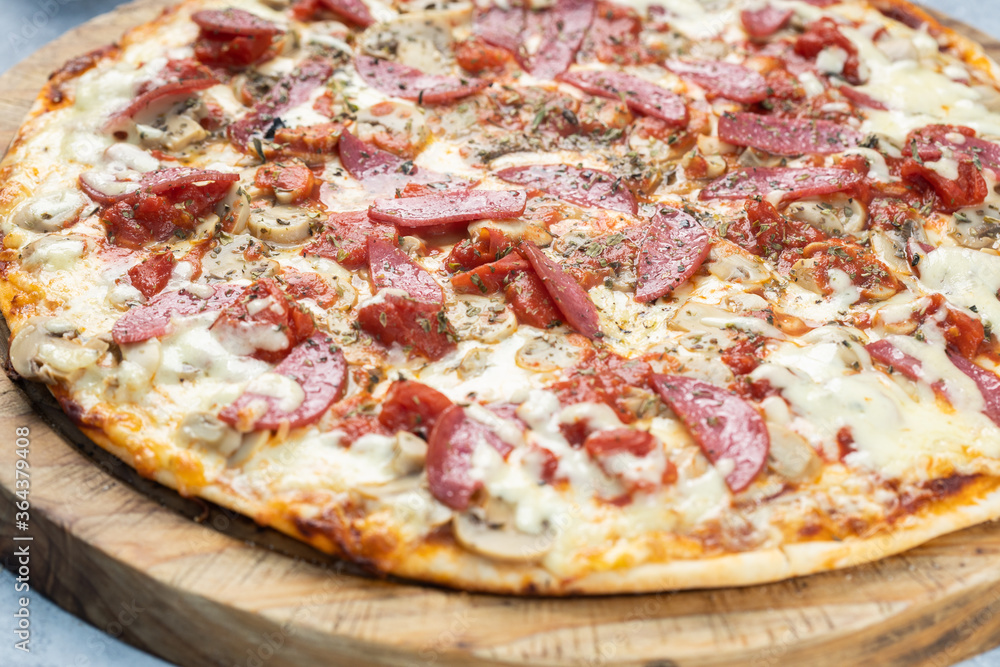 Close up of a pizza made in a wood-fired oven with salami and mushrooms, served on a rustic wooden board, isolated on a gray rustic textured wood.
