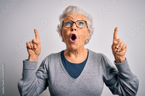 Senior beautiful grey-haired woman wearing casual sweater and glasses over white background amazed and surprised looking up and pointing with fingers and raised arms.