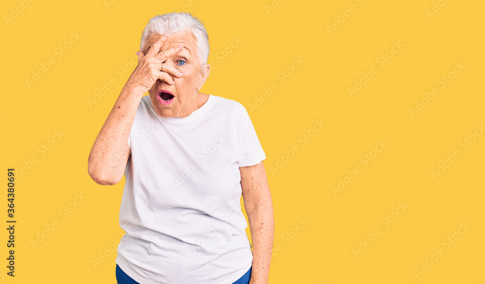 Senior beautiful woman with blue eyes and grey hair wearing casual white tshirt peeking in shock covering face and eyes with hand, looking through fingers with embarrassed expression. Senior beautiful woman with blue eyes and grey hair wearing casual white tshirt peeking in shock covering face and eyes with hand, looking through fingers with embarrassed expression.