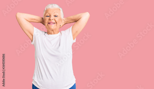Senior beautiful woman with blue eyes and grey hair wearing casual white tshirt relaxing and stretching, arms and hands behind head and neck smiling happy
