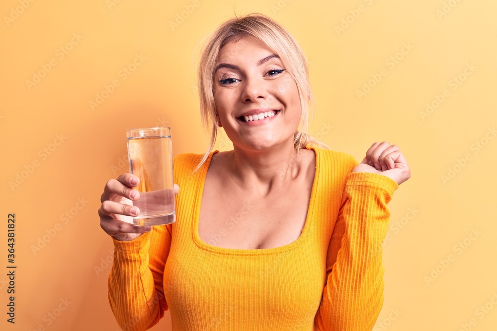 Young beautiful blonde woman drinking glass of water over isolated yellow background screaming proud, celebrating victory and success very excited with raised arm Young beautiful blonde woman drinking glass of water over isolated yellow background screaming proud, celebrating victory and success very excited with raised arm