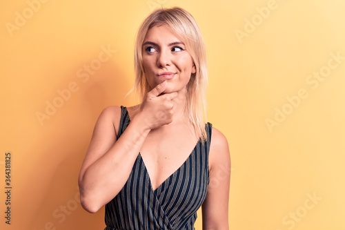 Young beautiful blonde woman wearing striped t-shirt standing over isolated yellow background thinking concentrated about doubt with finger on chin and looking up wondering