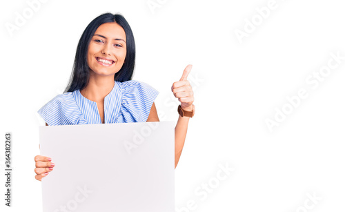 Young beautiful latin girl holding blank empty banner smiling happy and positive, thumb up doing excellent and approval sign