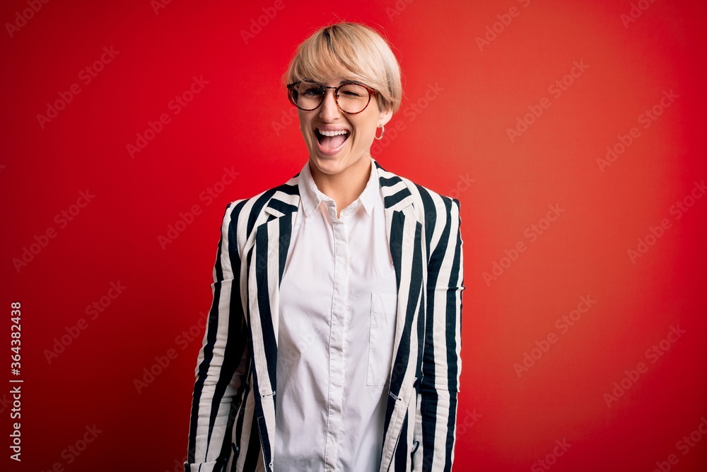 Blonde business woman with short hair wearing glasses and striped jacket over red background winking looking at the camera with sexy expression, cheerful and happy face. Blonde business woman with short hair wearing glasses and striped jacket over red background winking looking at the camera with sexy expression, cheerful and happy face.