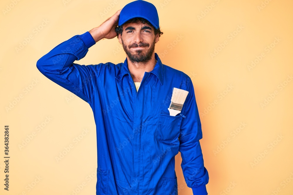Handsome young man with curly hair and bear wearing builder jumpsuit uniform confuse and wonder about question. uncertain with doubt, thinking with hand on head. pensive concept.