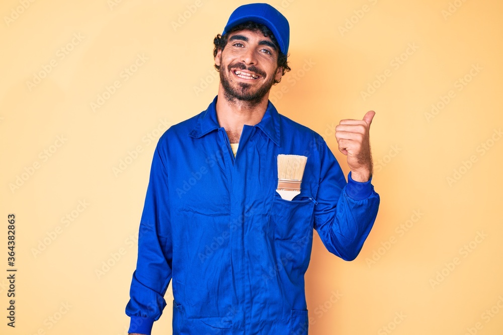Handsome young man with curly hair and bear wearing builder jumpsuit uniform smiling with happy face looking and pointing to the side with thumb up.