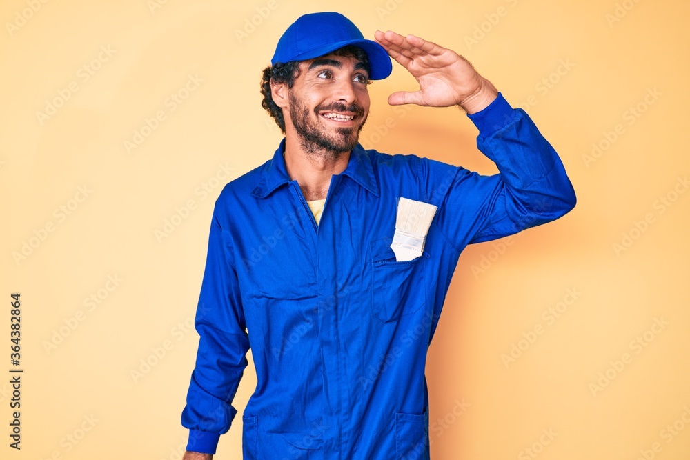 Handsome young man with curly hair and bear wearing builder jumpsuit uniform very happy and smiling looking far away with hand over head. searching concept.