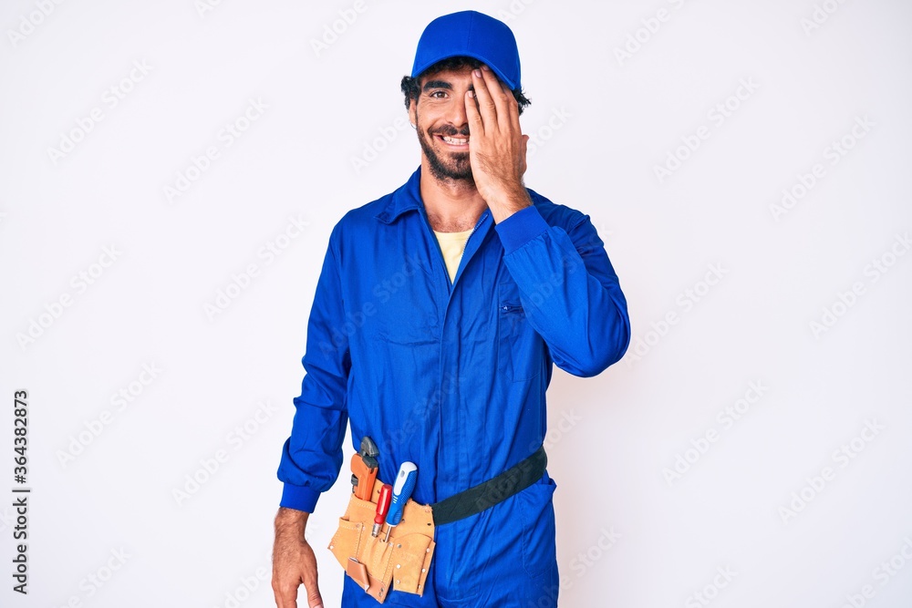 Handsome young man with curly hair and bear weaing handyman uniform covering one eye with hand, confident smile on face and surprise emotion.