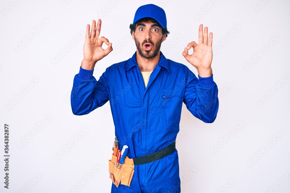Handsome young man with curly hair and bear weaing handyman uniform looking surprised and shocked doing ok approval symbol with fingers. crazy expression
