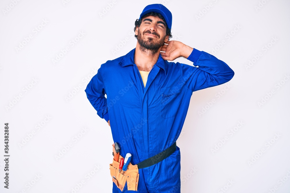 Handsome young man with curly hair and bear weaing handyman uniform suffering of neck ache injury, touching neck with hand, muscular pain Handsome young man with curly hair and bear weaing handyman uniform suffering of neck ache injury, touching neck with hand, muscular pain