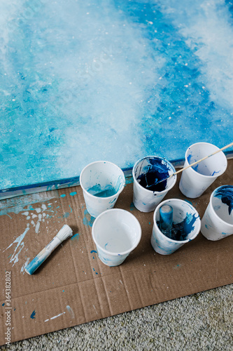 arts and craft hobbies concept, blue and white acrylic painting with colors in plastic cups and mixed painting tools next to it