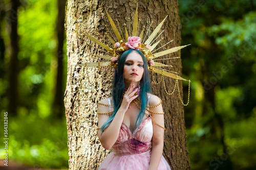 Costume Play. Magnificent Crowned Forest Nymph with Flowery Golden Crown Posing in Summer Forest Against High Tree Stem.