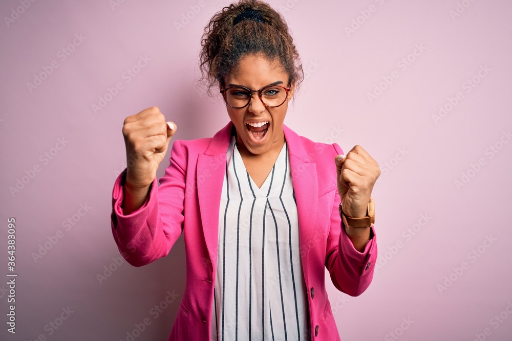 Beautiful african american businesswoman wearing jacket and glasses over pink background angry and mad raising fists frustrated and furious while shouting with anger. Rage and aggressive concept. Beautiful african american businesswoman wearing jacket and glasses over pink background angry and mad raising fists frustrated and furious while shouting with anger. Rage and aggressive concept.