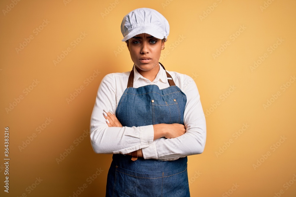 Young african american afro baker woman wearing apron and cap over yellow background skeptic and nervous, disapproving expression on face with crossed arms. Negative person.