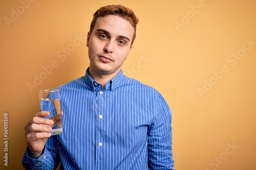 Young handsome redhead man drinking glass of water over isolated yellow background thinking attitude and sober expression looking self confident © Krakenimages.com