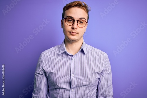 Young handsome redhead man wearing casual shirt and glasses over purple background Relaxed with serious expression on face. Simple and natural looking at the camera. © Krakenimages.com