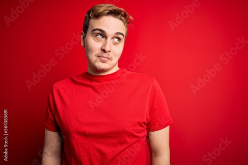 Young handsome redhead man wearing casual t-shirt over isolated red background smiling looking to the side and staring away thinking.