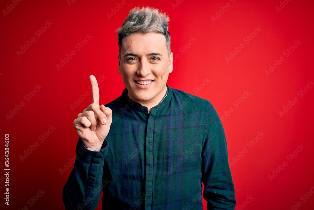 Young handsome modern man wearing elegant green shirt over red isolated background showing and pointing up with finger number one while smiling confident and happy. Young handsome modern man wearing elegant green shirt over red isolated background showing and pointing up with finger number one while smiling confident and happy.