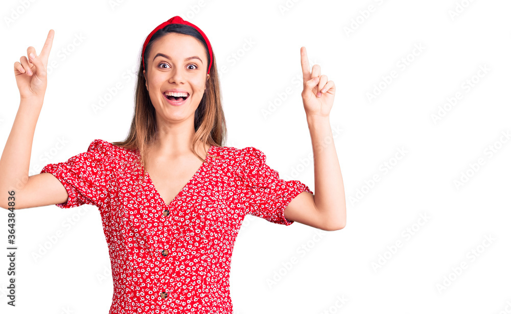 Young beautiful girl wearing dress and diadem smiling amazed and surprised and pointing up with fingers and raised arms. Young beautiful girl wearing dress and diadem smiling amazed and surprised and pointing up with fingers and raised arms.