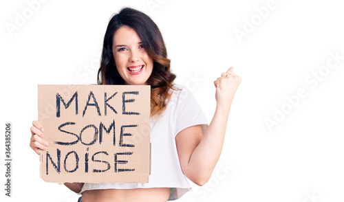 Beautiful young brunette woman holding make some noise banner screaming proud, celebrating victory and success very excited with raised arms photo